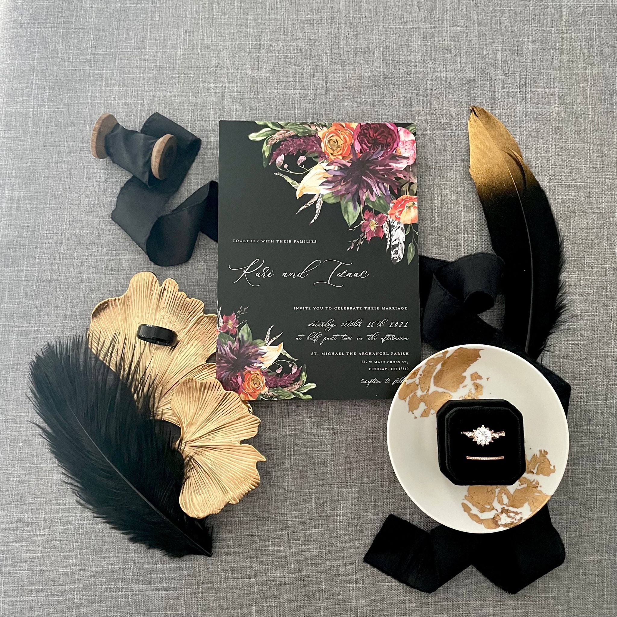 Black wedding invitation with deep pink, purple and orange flowers.  Black ring box is in a white dish with a black silk ribbon styled around it with a gold gingko leaf dish on one side and gold dipped feather on the other