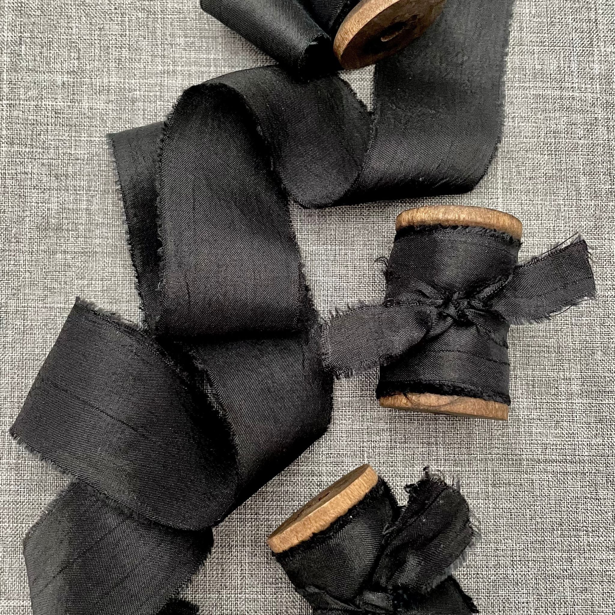 4 spools of black silk ribbon, 3 are rolled up and one is partially unrolled, they are styled on a mid tone gray background