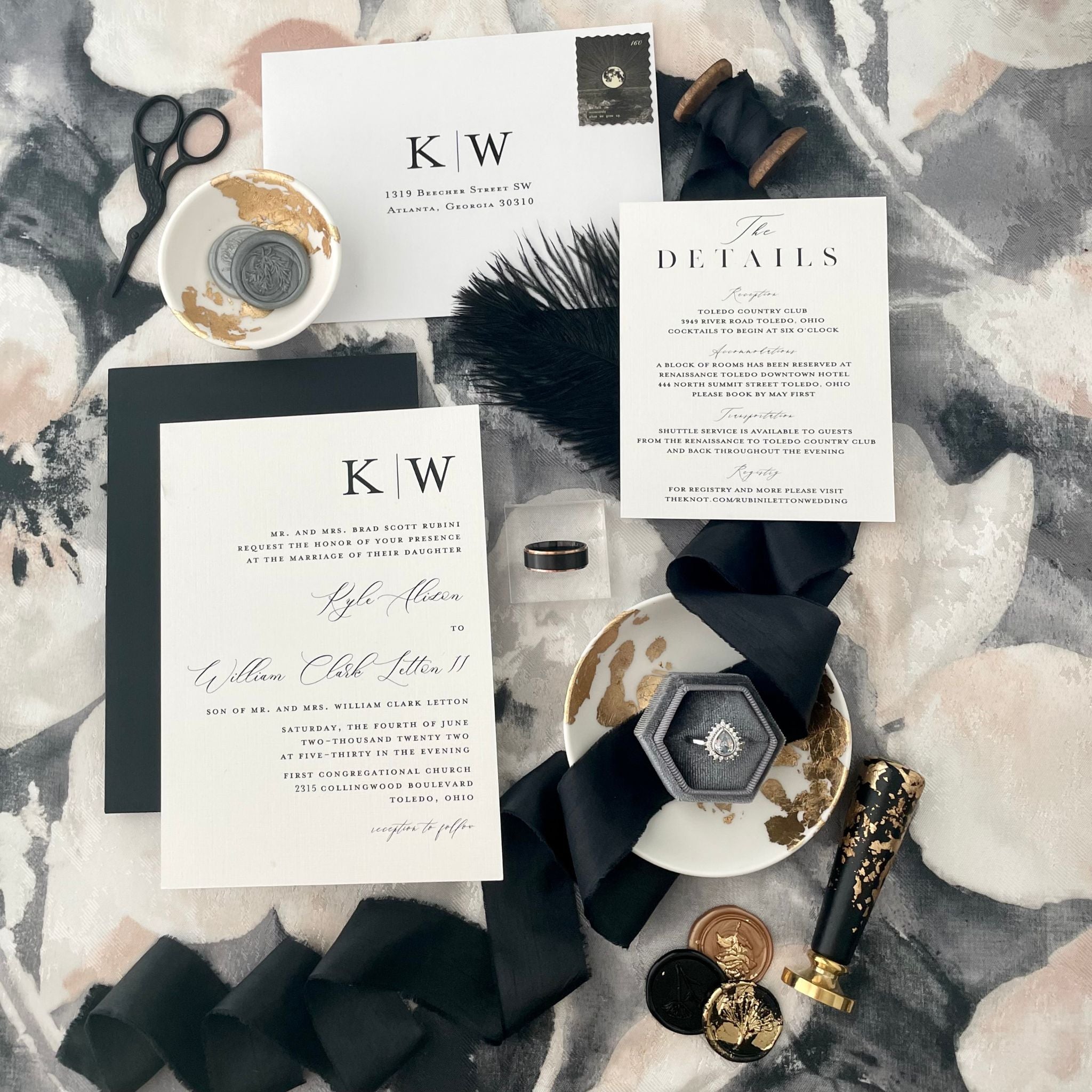 White invitation with black modern font on a black, white and gray floral background.  Flatlay details included a black silk ribbon unrolled with gray ring box and small white dish with wax seals.