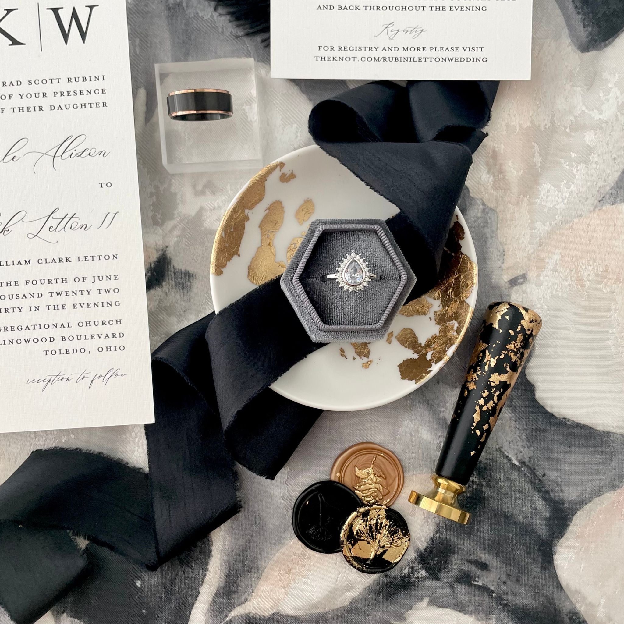 White ring dish with gold leafing with a gray hexagon ring box with a silk black ribbon styled throught the center, there are 3 wax seals in black and gold and wax stamper styled with it on a black white and gray floral background