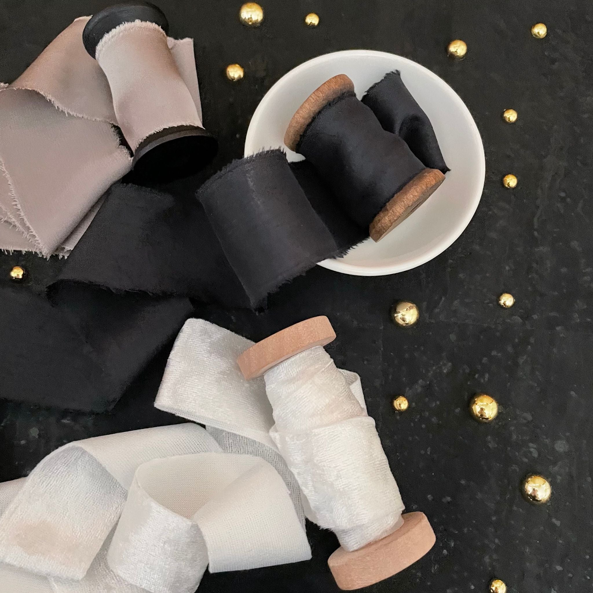 3 spools of ribbon slightly unrolled on a black background with small gold pearls.  The ribbon is a black silk, white velvet and light gray silk.
