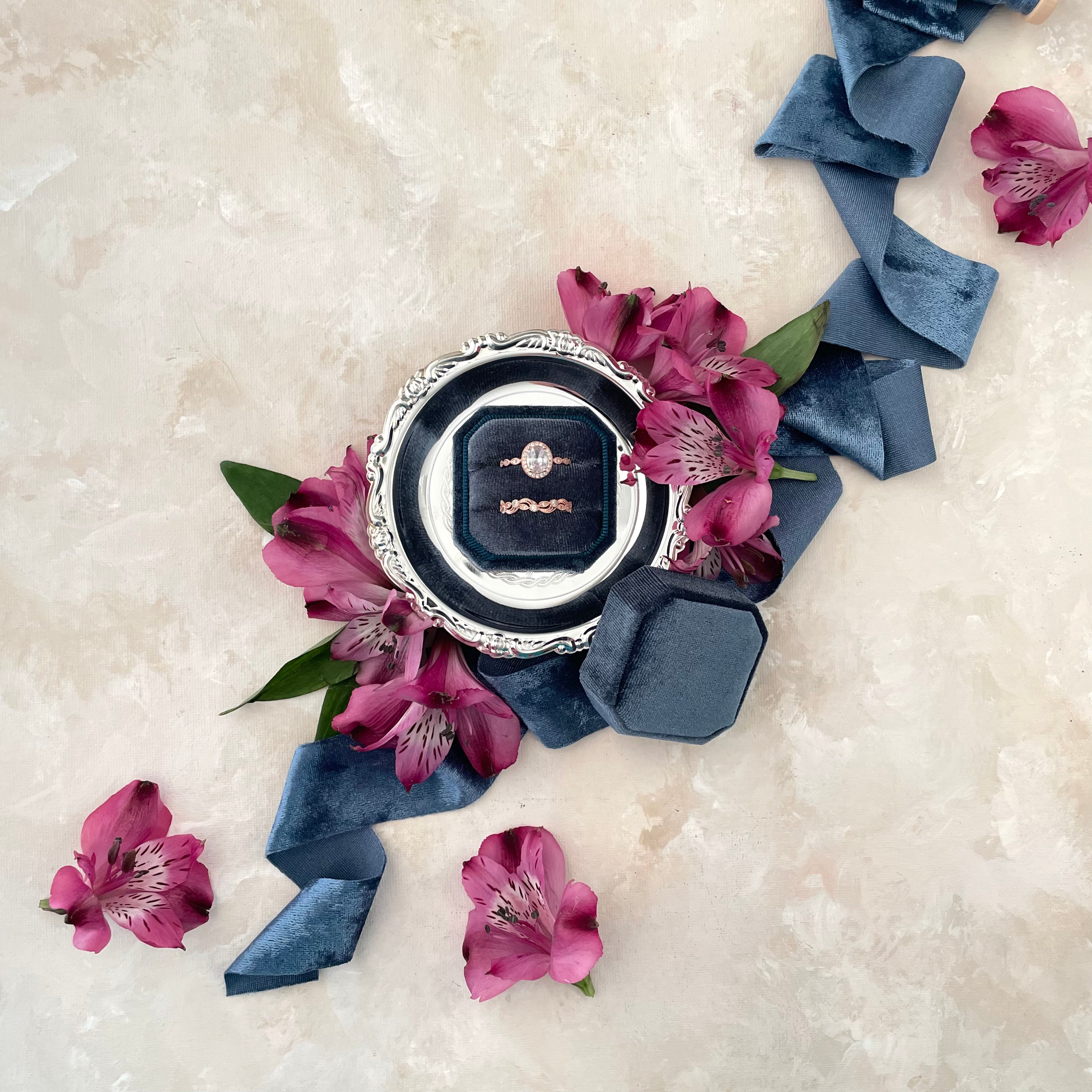 Navy blue ring box on silver round mini styling tray with blue velvet ribbon and purple flowers - Flat lay props from Champagne & GRIT