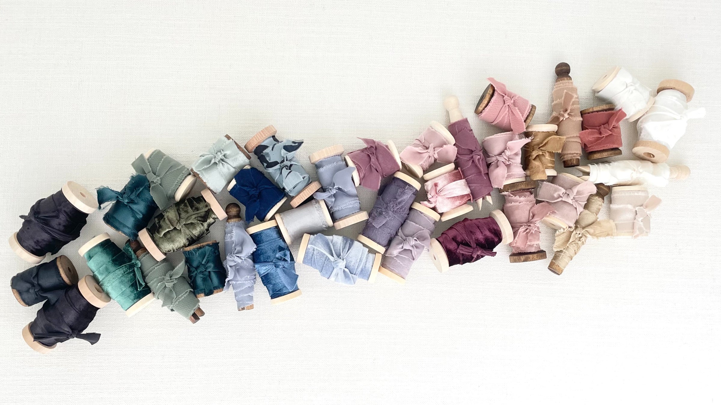 A variety of ribbon laid out in a row including the colors black, sage green, navy blue, dusty blue, lavender, dusty pink, tan, and ivory from Champagne & GRIT