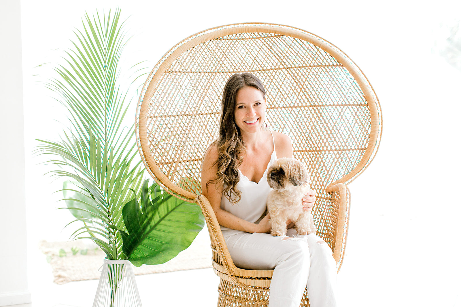 Jaime, owner of Champagne & GRIT smiling while sitting in a wicker chair with her dog on her lap