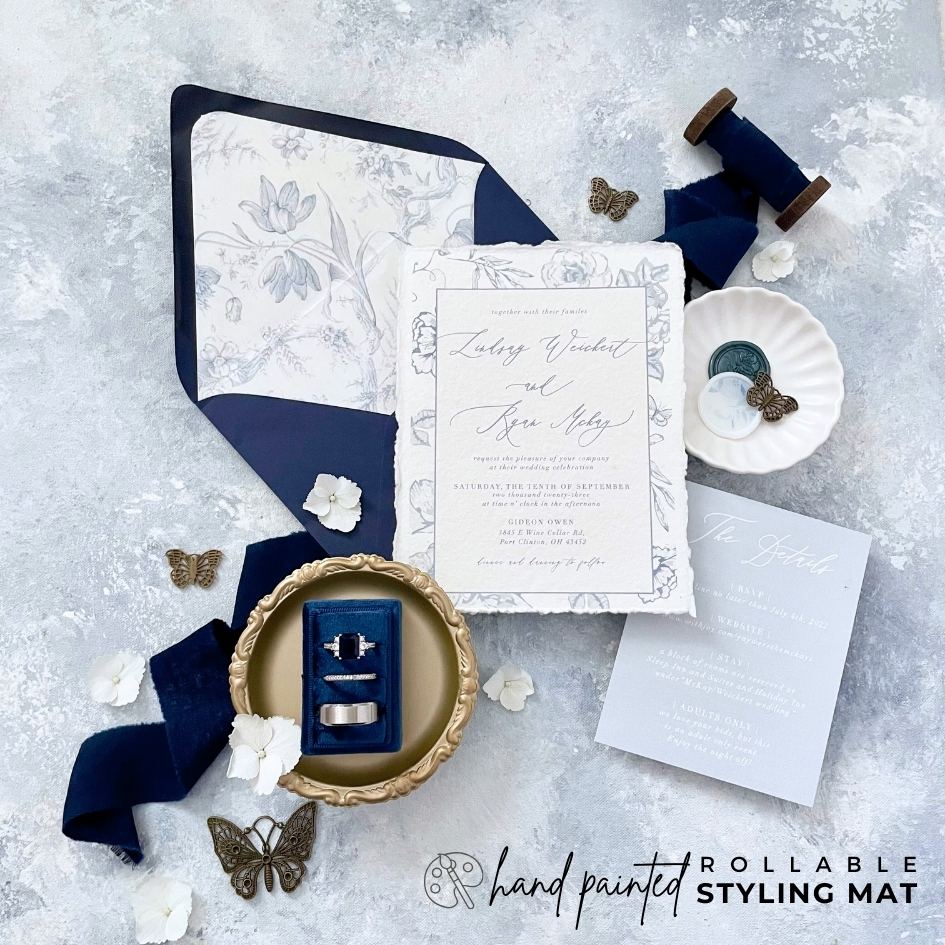 Navy Blue Floral Invitation Suite Flat Lay Styled on Dusty Blue Background.  Rollable Dusty Blue Styling Mat Showcases white Ring Dish, Navy 3 slot ring box  silk Navy blue flatlay ribbon & Butterflies.