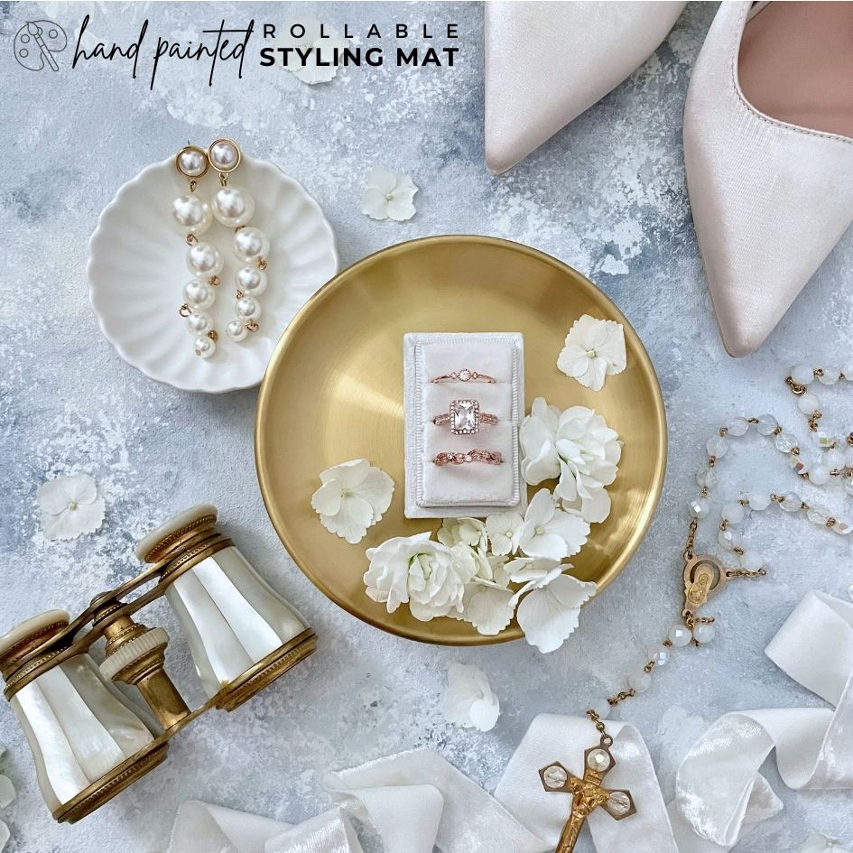 Bridal Flatlay detail showcasing, 3 slot ring box on a gold dish, ivory pumps, white velvet ribbon, a small white ring dish & bridal earrings.  Wedding flat lay is Styled on a Light blue flat lay Styling mat, that is Hand painted with shades of blue & touches of white.