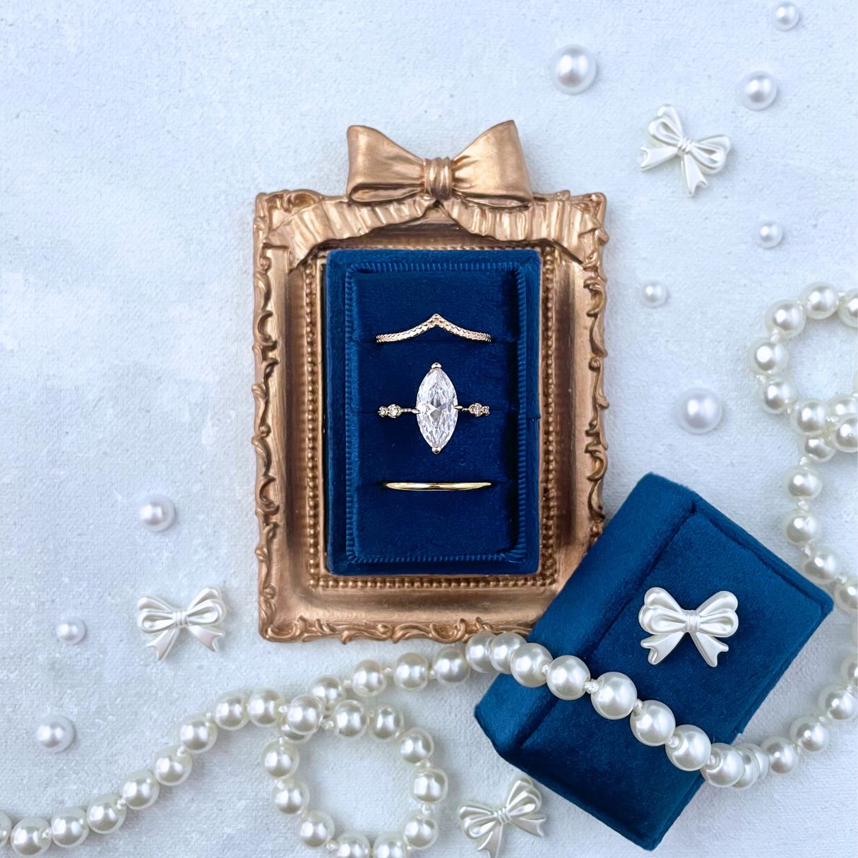 Navy Blue Ring box with 3 slots set inside a mini Gold Frame with bow onto.  Styled with pearls, and bows on a light blue background