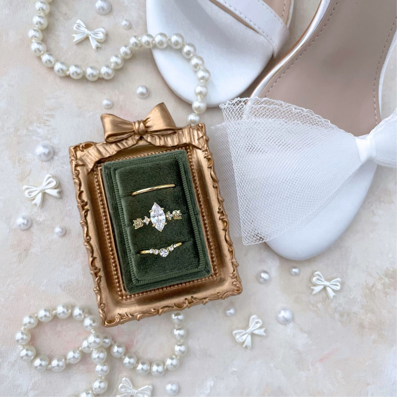 mini Gold frame with a bow ontop with an olive green velvet rectangle ring box.  Ring Box has 3 gold rings with diamonds. Styled with a pearl necklace, and white satin shoeswith bow on a light background.