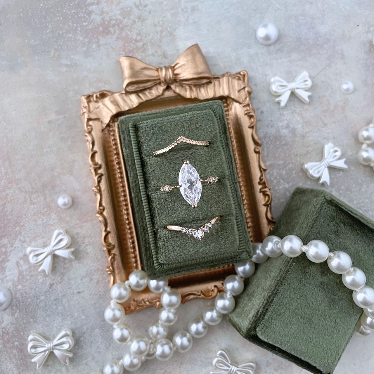 mini Gold frame with a bow onto.  inside the frame is an olive green ring box with 3 gold rings.  Styled with a white pearl necklace & mini pearlized bow & loose pearls. 