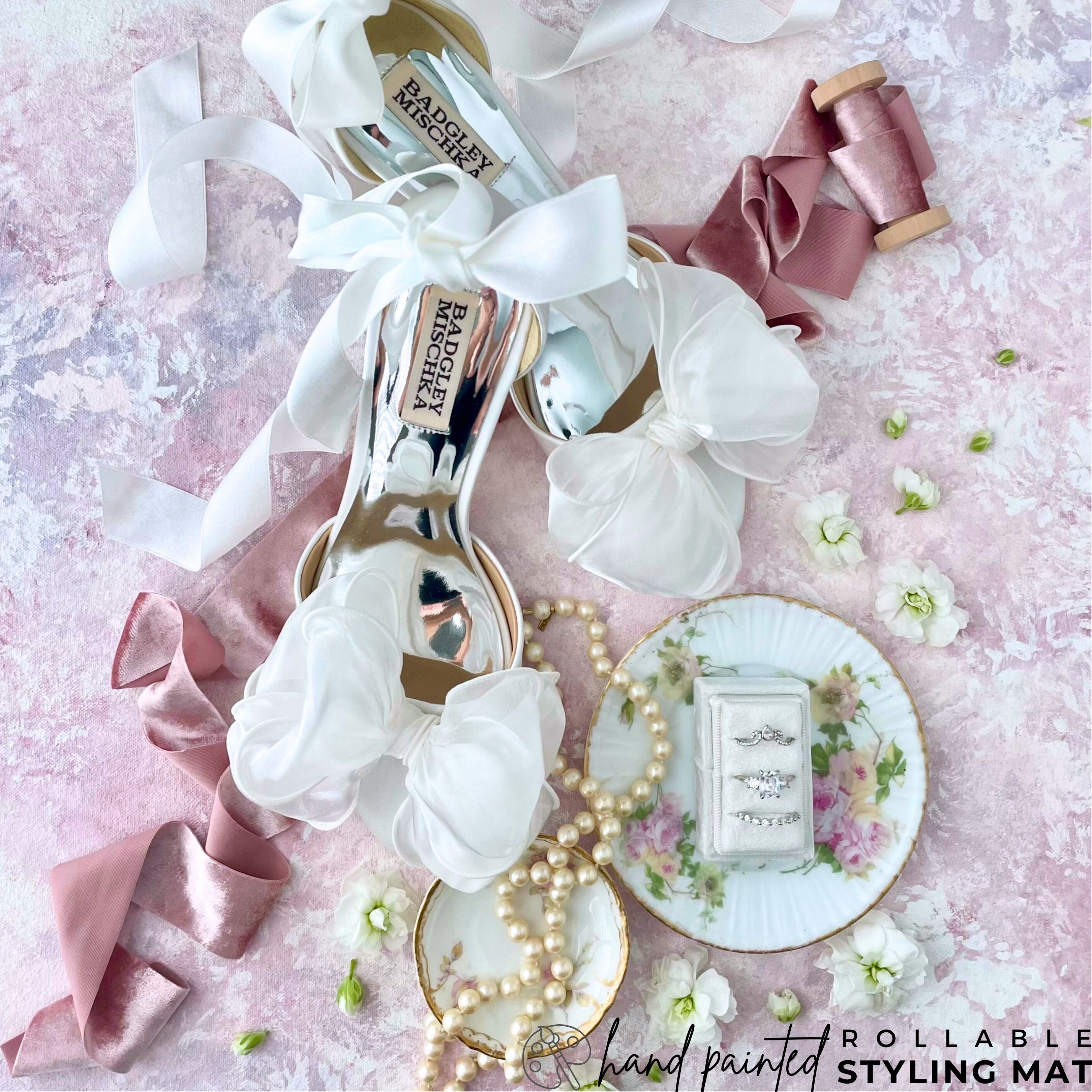 Light Pink & Purple Flat Lay Styling Mat showcases a Bridal detail, Featuring Silk Bow Pumps.  Shoes are Styled on Pink Hand Painted Backdrop with White flowers & Vintage ring dish.  Text on image; hand painted rollable Styling mat