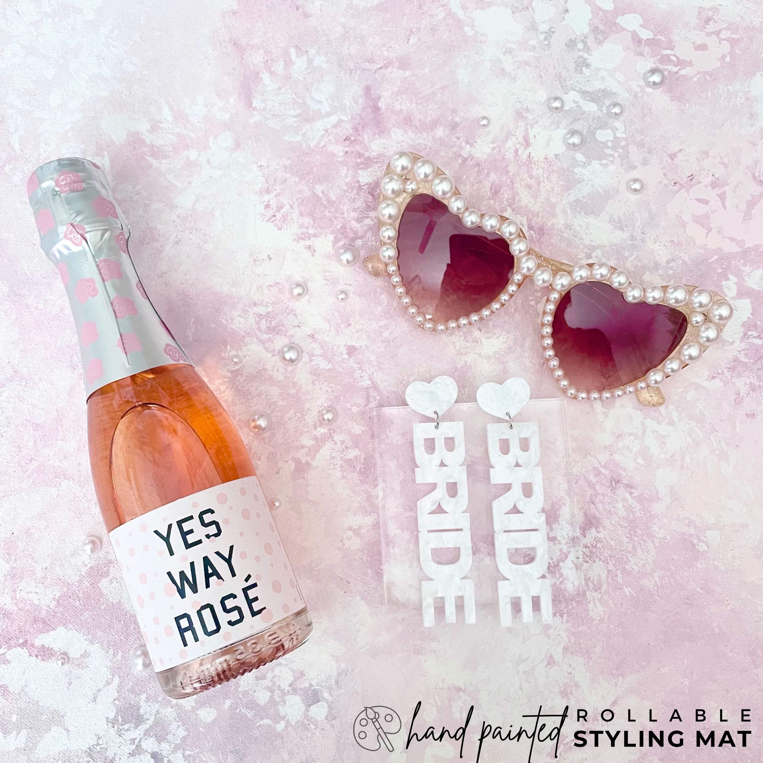 Bridal Flatlay detail showcasing, heart shaped sunglasses, a mini champagne Bottle & Bride Earrings.  Wedding Flat Lay is Styled on a Hand painted lavender & Mauve FlatLay Styling Mat 