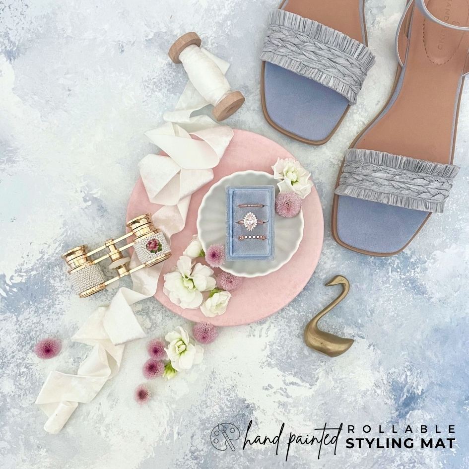 Dusty Blue Bridal Flat lay showcasing, Dusty Blue 3 slot ring box on a white ring dish, light blue sandals, vintage pearl opera glasses, white velvet ribbon.  Wedding flatlay is Styled on a dusty blue flat lay Styling mat, that is rollable & Hand painted with shades of blue & touches of white.