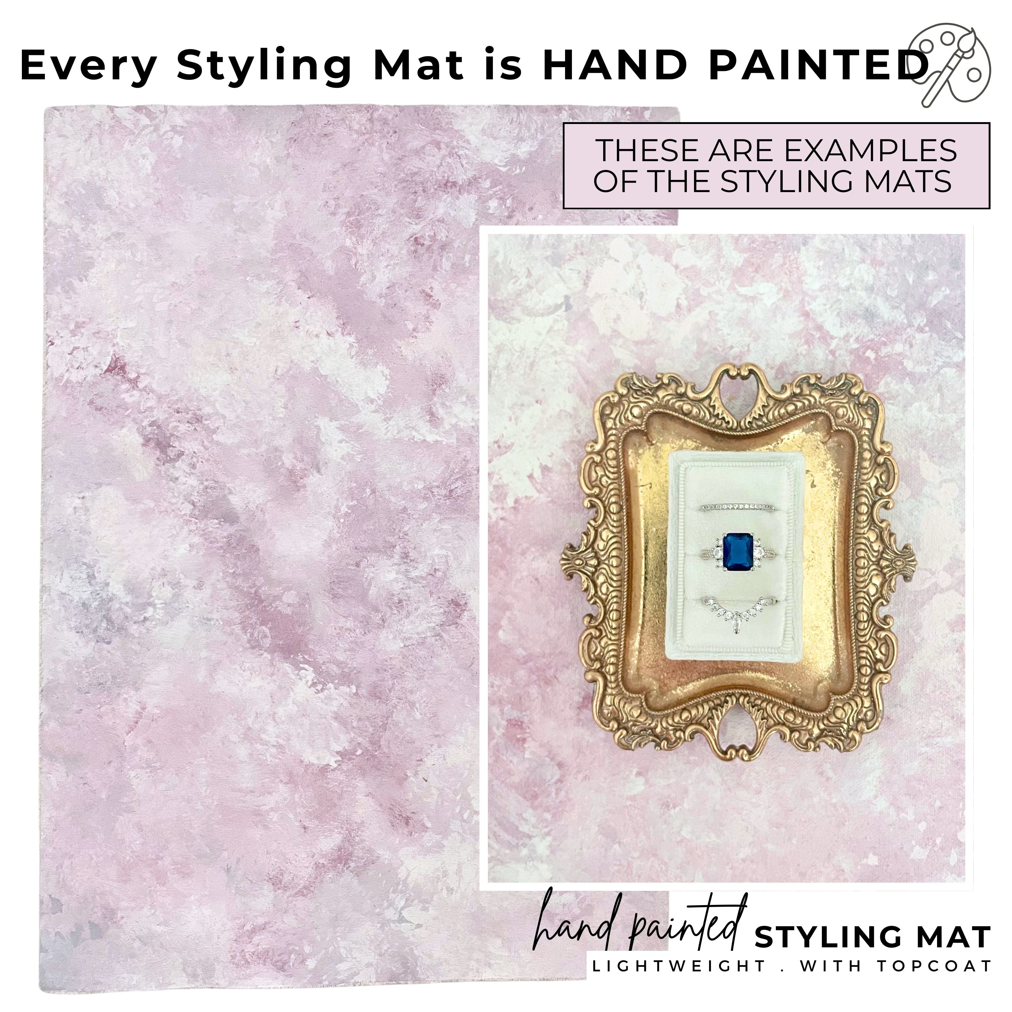 Text says: Every Flat Lay Styling mat is hand painted.  Rollable styling surface in dusty rose & lilac color pallet for photographers.  Ring Detail showcased, mini gold tray with white 3 slot ring box styled on a Pink & purple flatlay background.