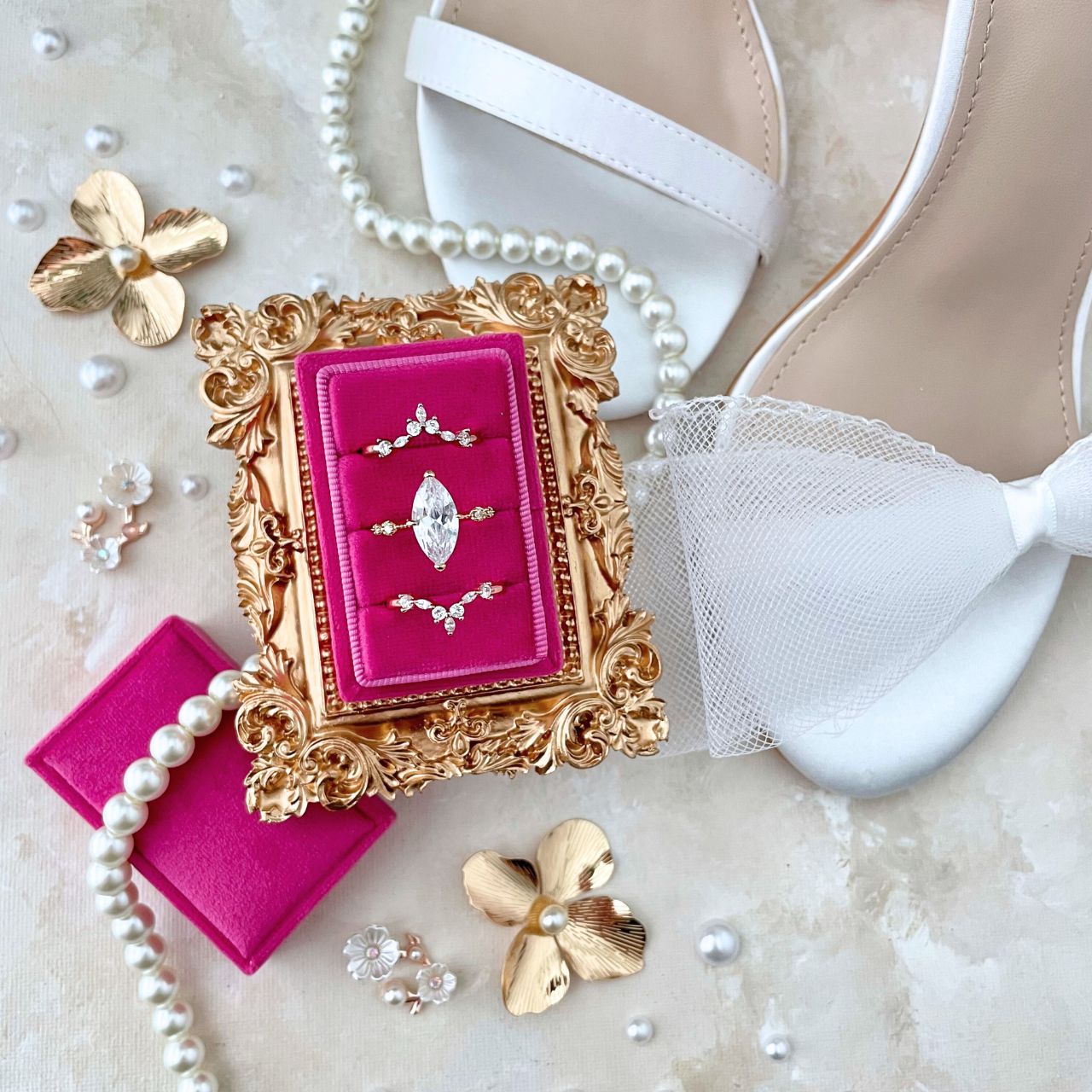 a bridal shoe flat lay detail of a hot pink rectangle ring box, with 3 rings.  Styled with a mini golden frame with a bow on top, white shoes with bows on the strap, a pearl necklace and golden flowers and pearls.  This flat lay is on a hand-painted neutral canvas of tans and white with touches of pink.
