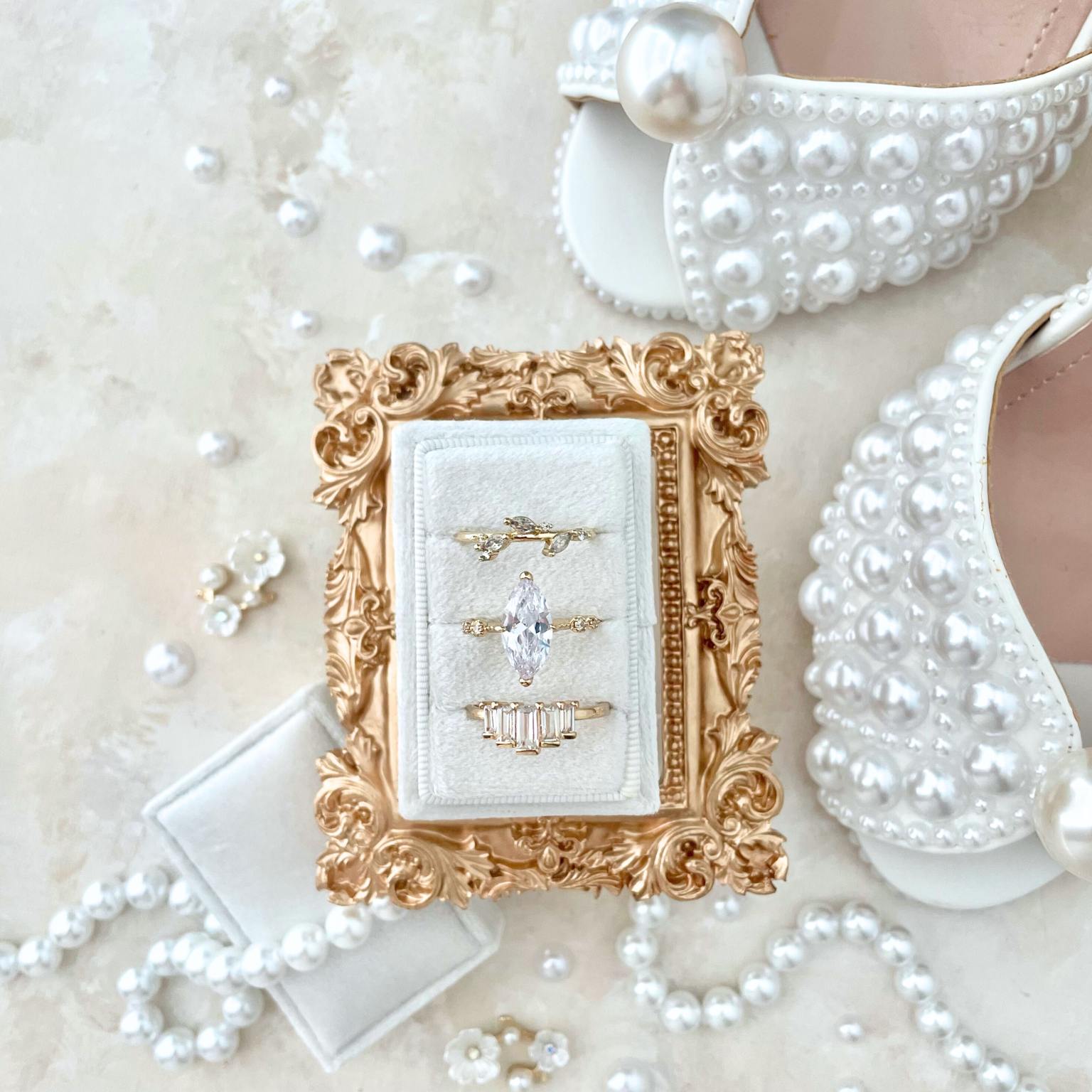 White rectangle ring box with 3 gold rings.  Ring box is set inside a vintage inspired gold frame.  Styled on a beige background with pearls and white shoes.