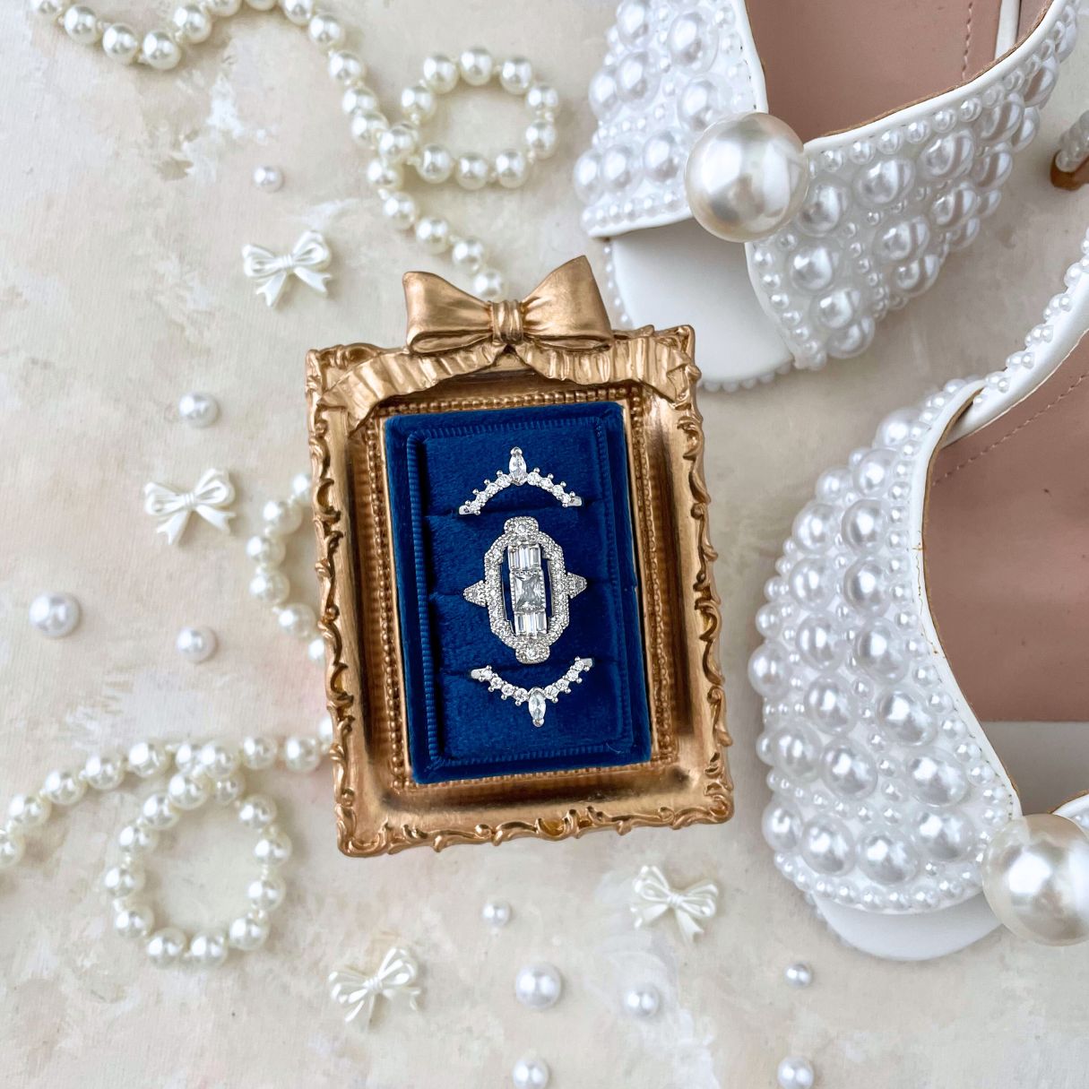Wedding Flat lay detail of a Navy Blue rectangle ring box, with 3 rings.  Styled with a mini golden frame with a bow on top, pearl shoes, a pearl necklace and mini bows and pearls.  This flat lay is on a neutral styling canvas in shades of tan and white.
