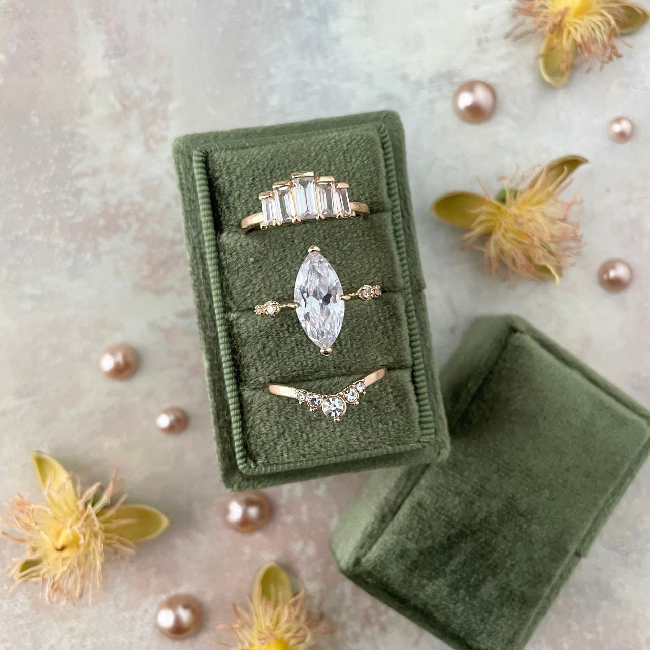 Set of gold rings with a central diamond in a  slot olive green velvet box, surrounded by pearls and flowers on a light background.