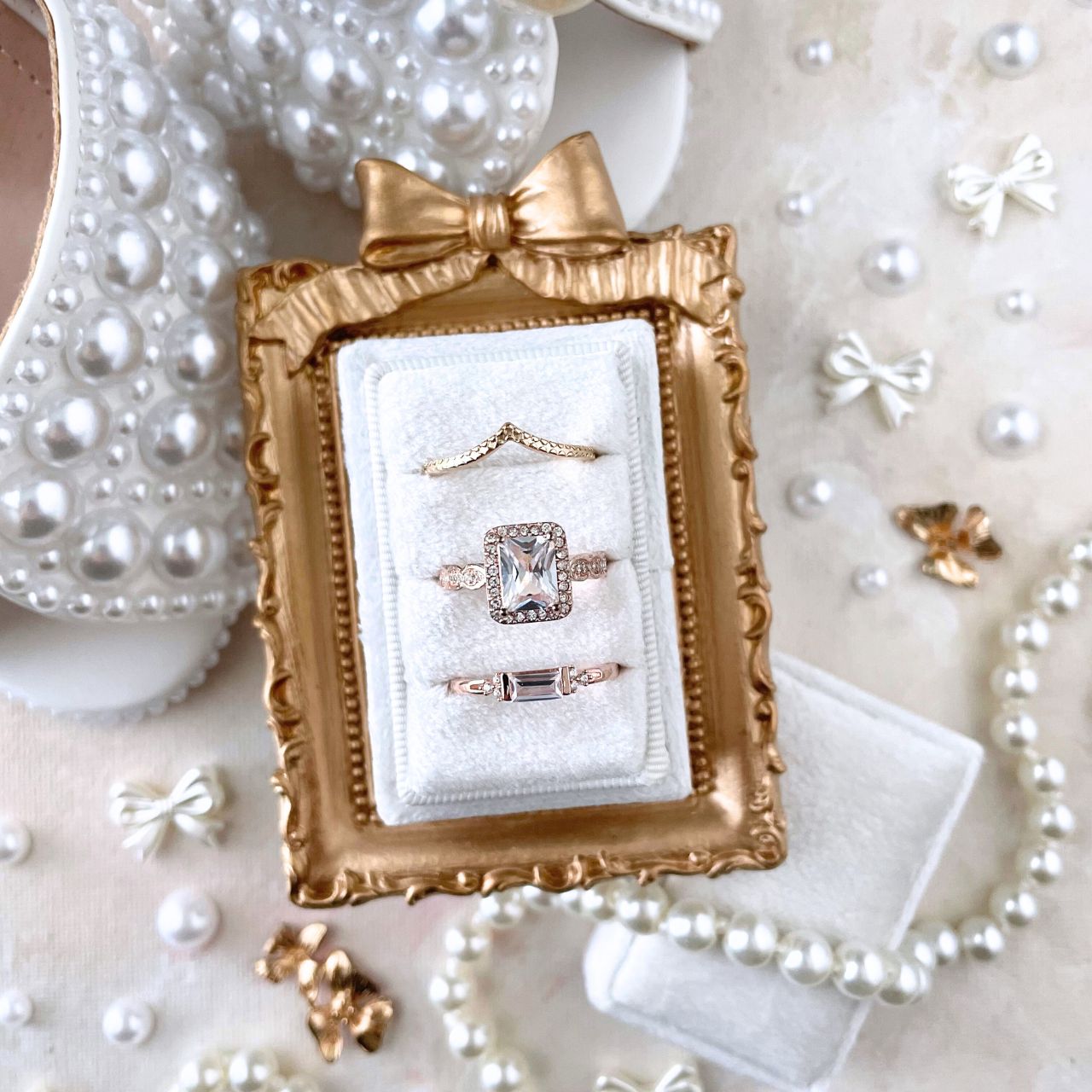 Wedding Flat lay detail of a white velvet rectangle ring box, with 3 gold rings.  Styled with a mini golden frame with a bow on top, pearl shoes, a pearl necklace and mini bows and pearls.  This flat lay is on a neutral styling canvas in shades of tan and white.