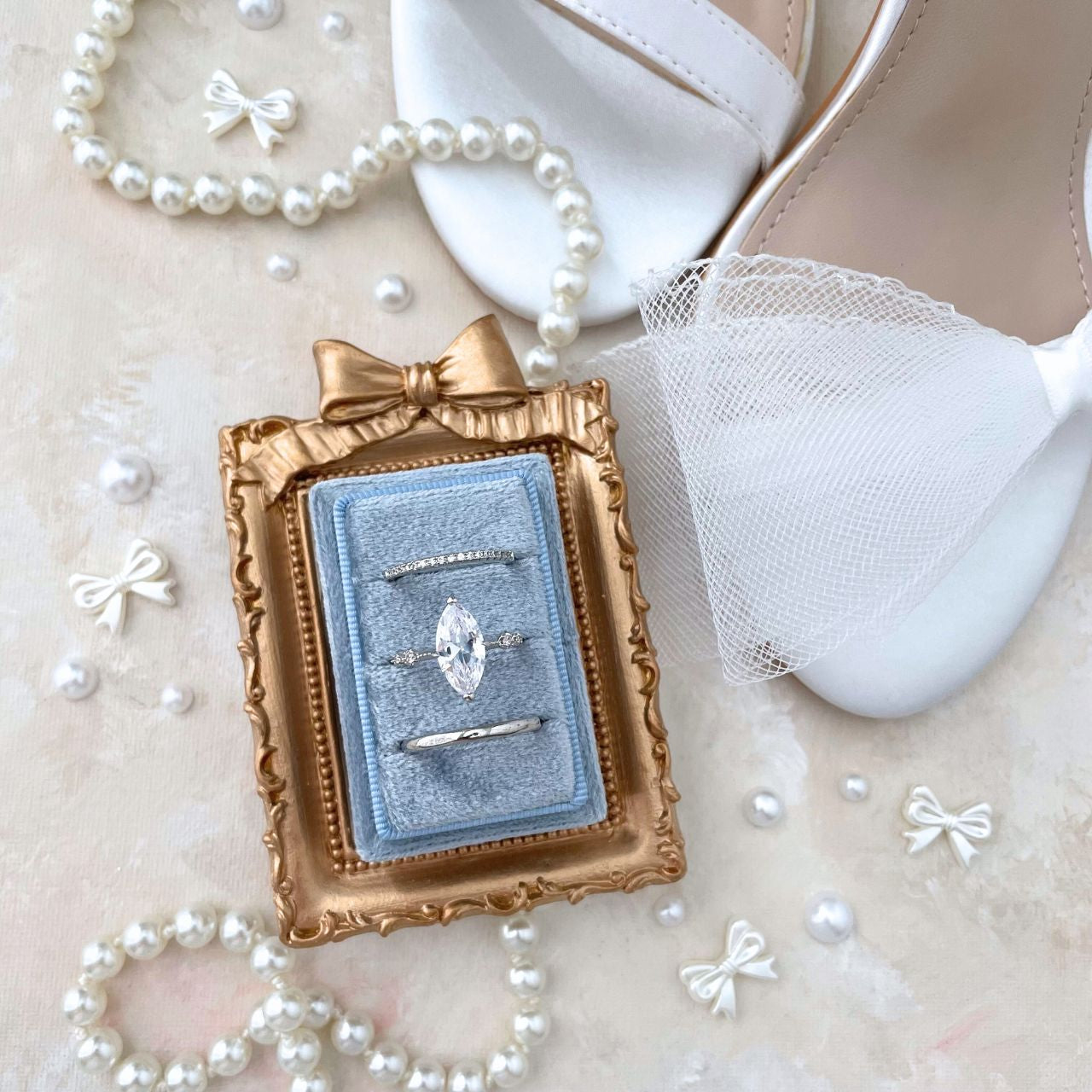Mini Gold frame with a bow onto,  Centered inside is a dusty blue ring box.  Ring box has 3 slots and is rectangles.  Styled with  a pearl necklace & loose mini bows and pearls.  