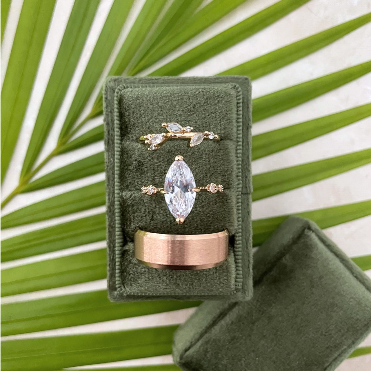 Olive Green Velvet Rectangle ring box, with 3 slots, with 3 gold rings.  styled onto of  palm leaf