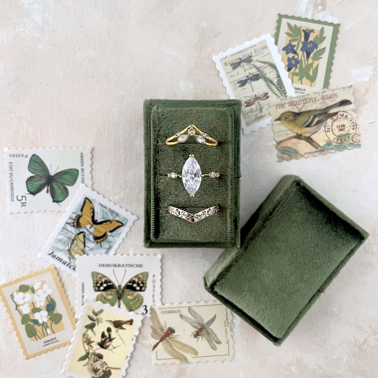 Olive green Ring Holder with 3 slots with 3 gold rings.  Ring Box is  surrounded by vintage-style postage stamps featuring nature-themed designs, in shades of greens