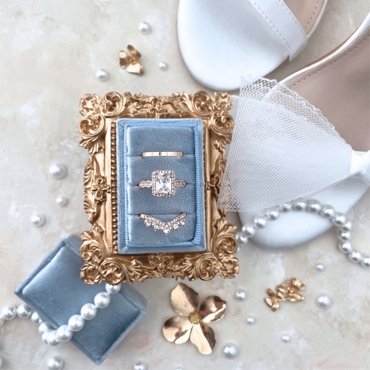 a bridal shoe flat lay detail of a dusty blue velvet rectangle ring box, with 3 gold rings.  Styled with a mini golden vintage inspired frame with.  Silk white bridal shoes have a bow on the strap.   Styled with a  pearl necklace and mini golden flowers and pearls.  This flat lay is on a hand-painted neutral canvas of tans and white with touches of pink.