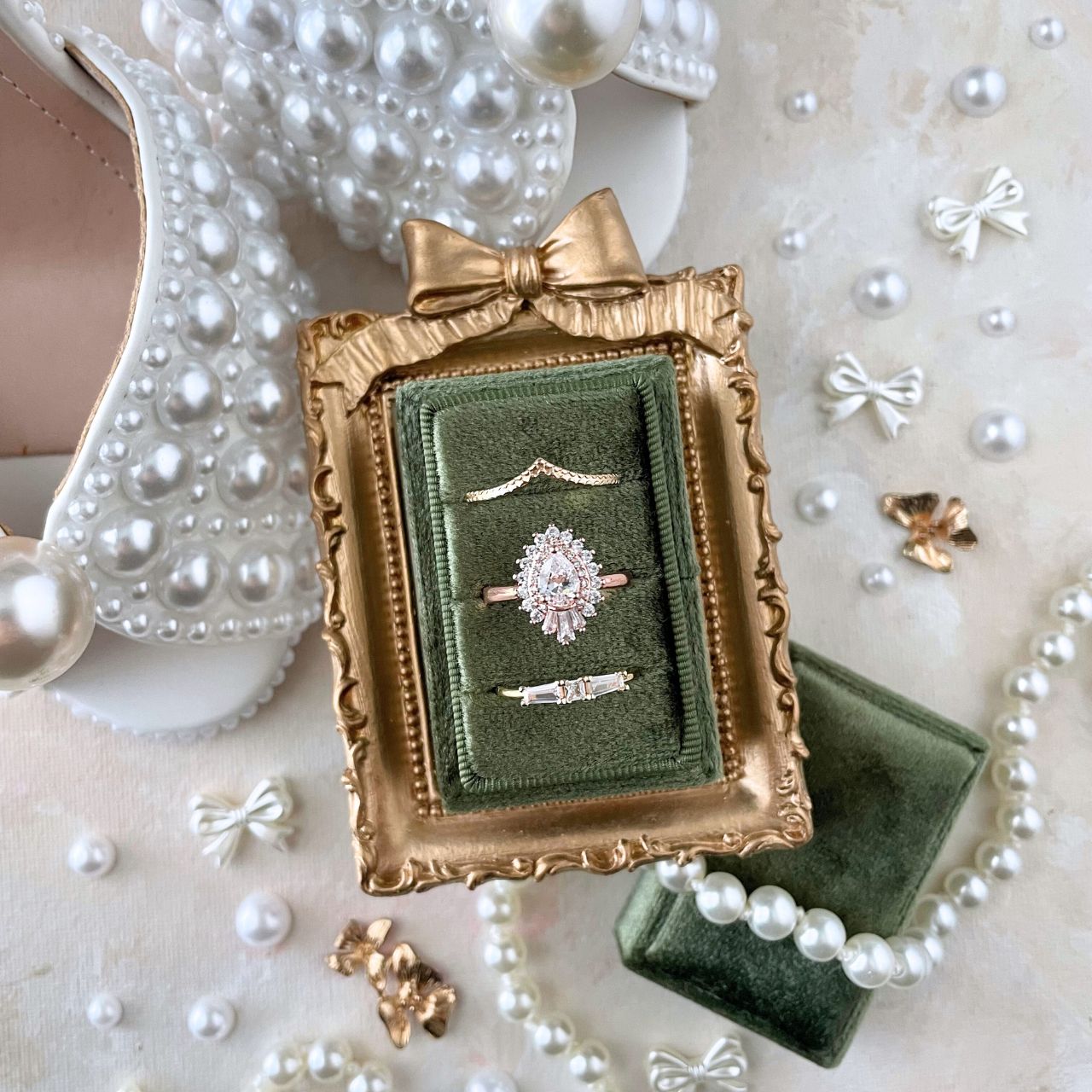 Gold mini frame with a bow onto, showcasing an olive green 3 slot ring box with 3 gold diamond rings. surrounded by pearls and decorative bows.