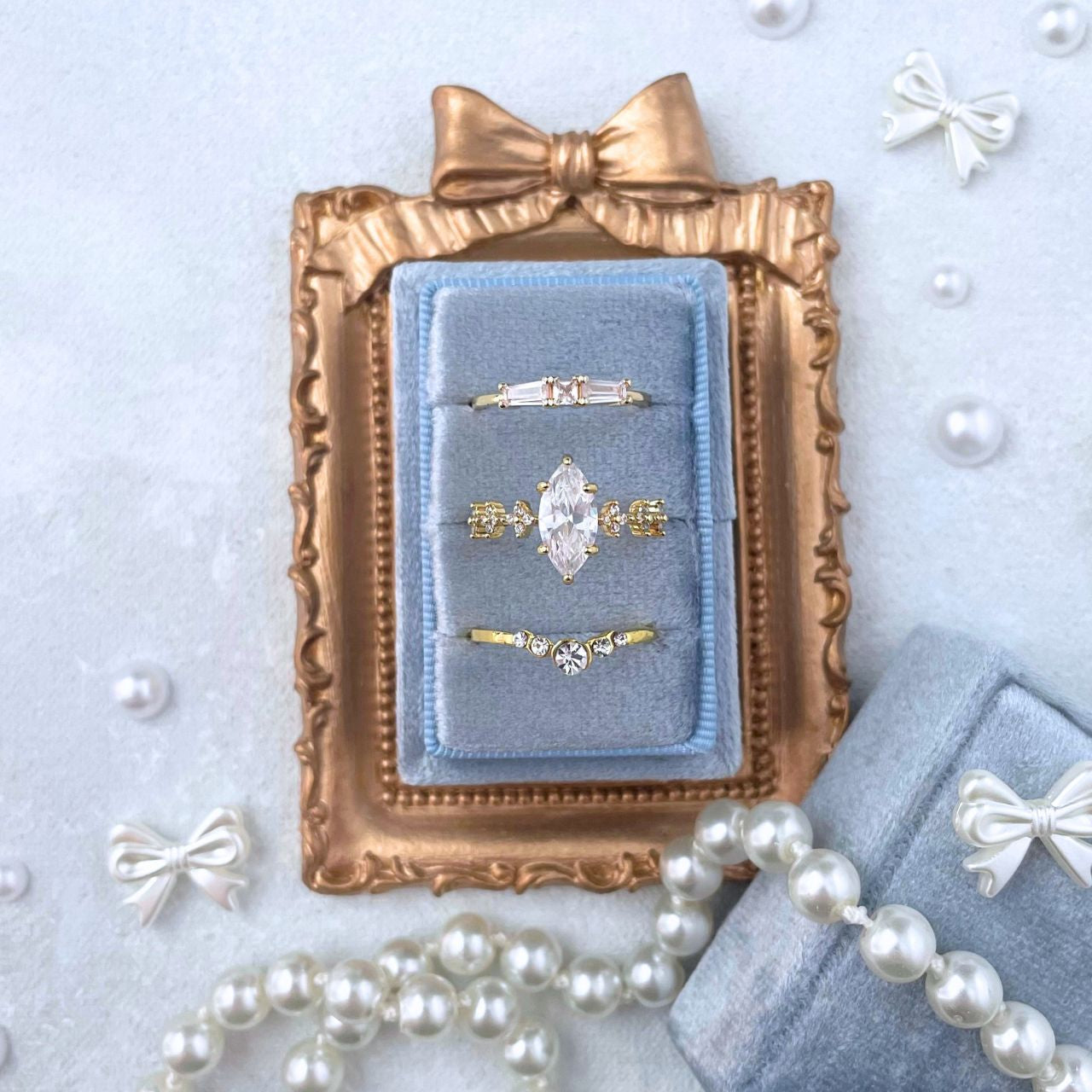 Gold. mini fram with a bow onto.  Set inside is a dusty blue 3 slot velvet ring box .  Ring Box is holding 3 golden rings.  Styled with jmini pearlized white bows, surrounded by pearls on a light blue background