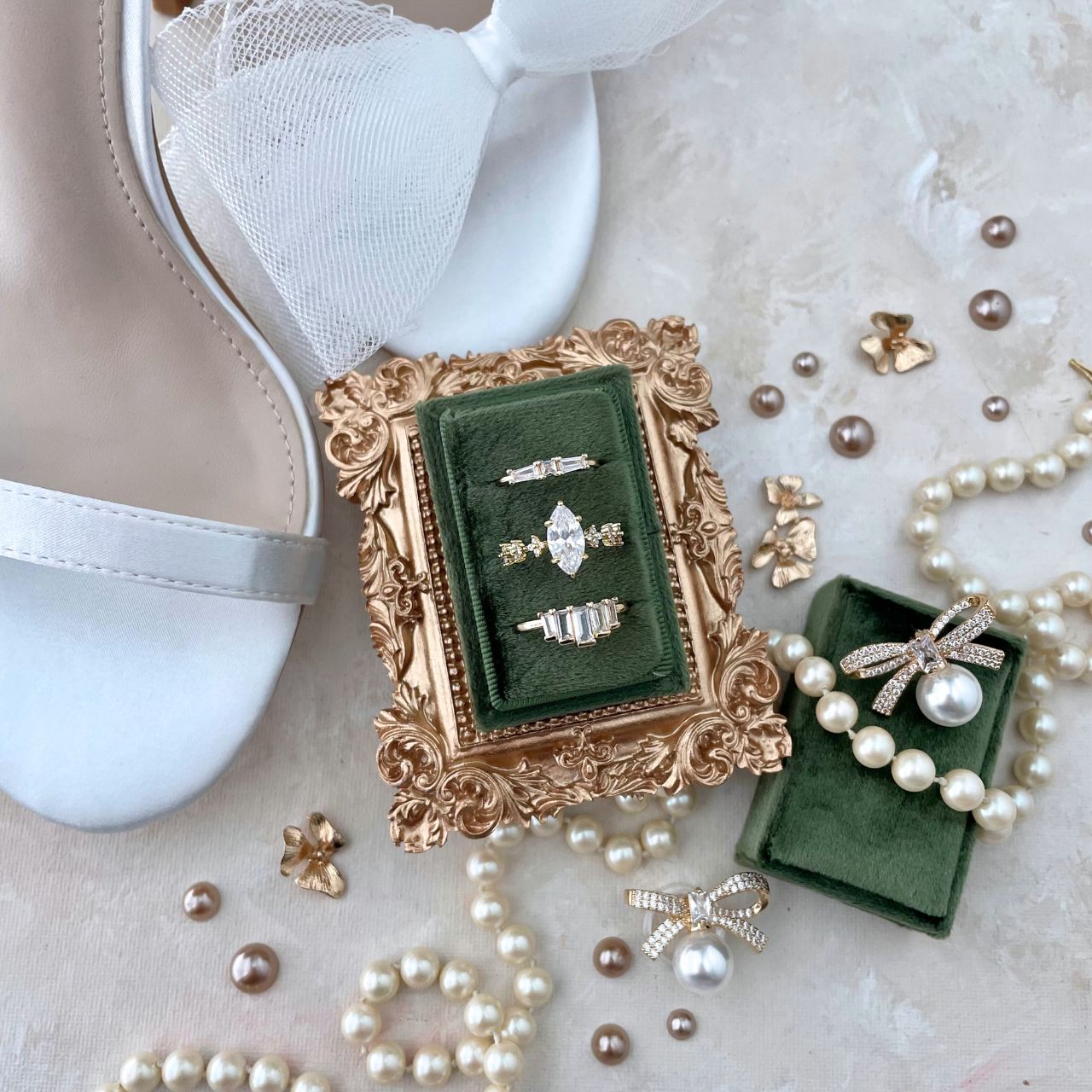 Wedding Flat lay detail of a olive green rectangle ring box, with 3 gold rings.  Styled with a mini golden frame vintage style frame.  Styled with champagne pearls & a pearl necklace.  There is a white silk pair of shoes with a bow on strap. This flat lay is on a neutral styling canvas in shades of tan and white.
