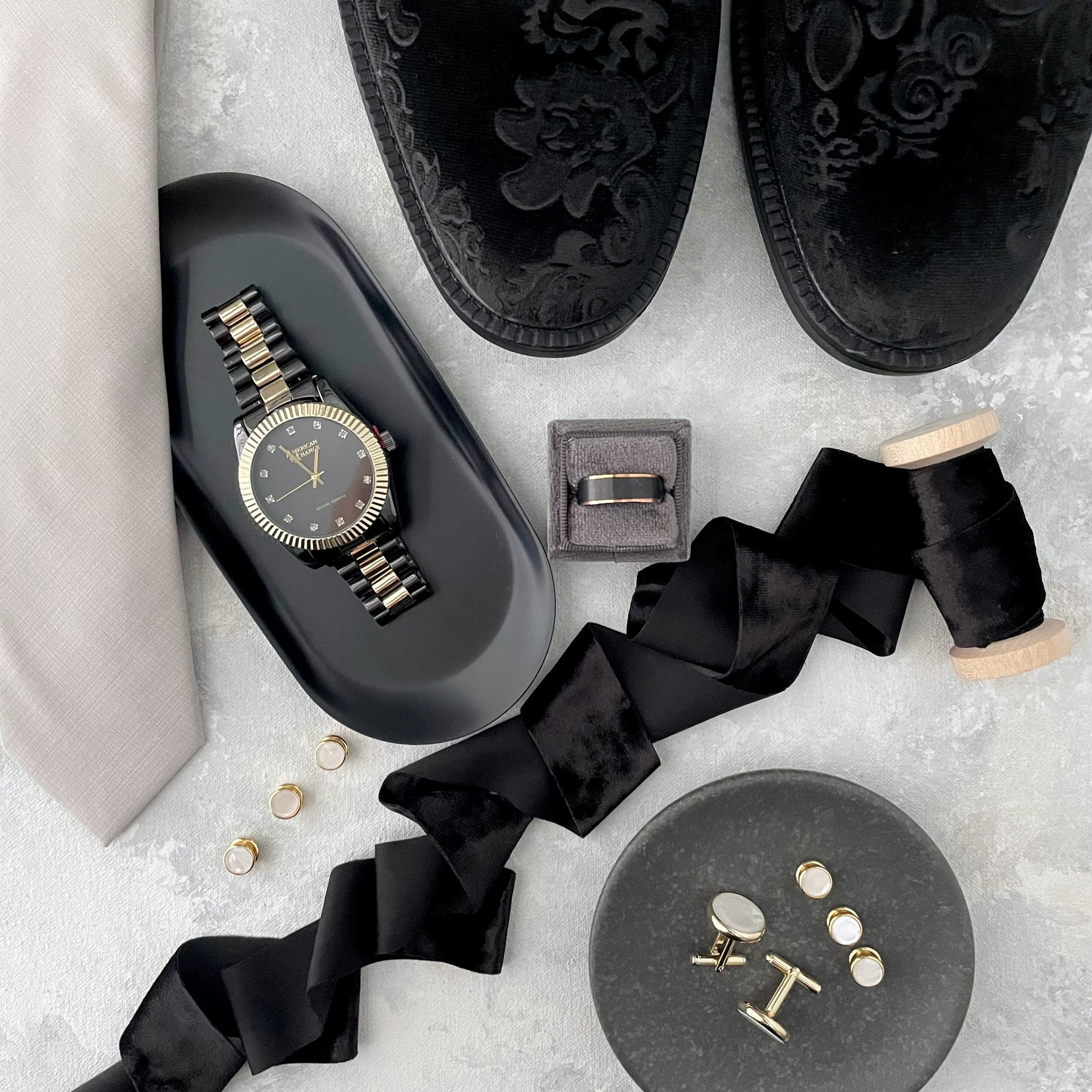 Black velvet mens shoes, with cufflinks, black and gold watch on a black oval dish, tan tie & black velvet ribbon on wood spool.  Gray square ring box with black grooms ring