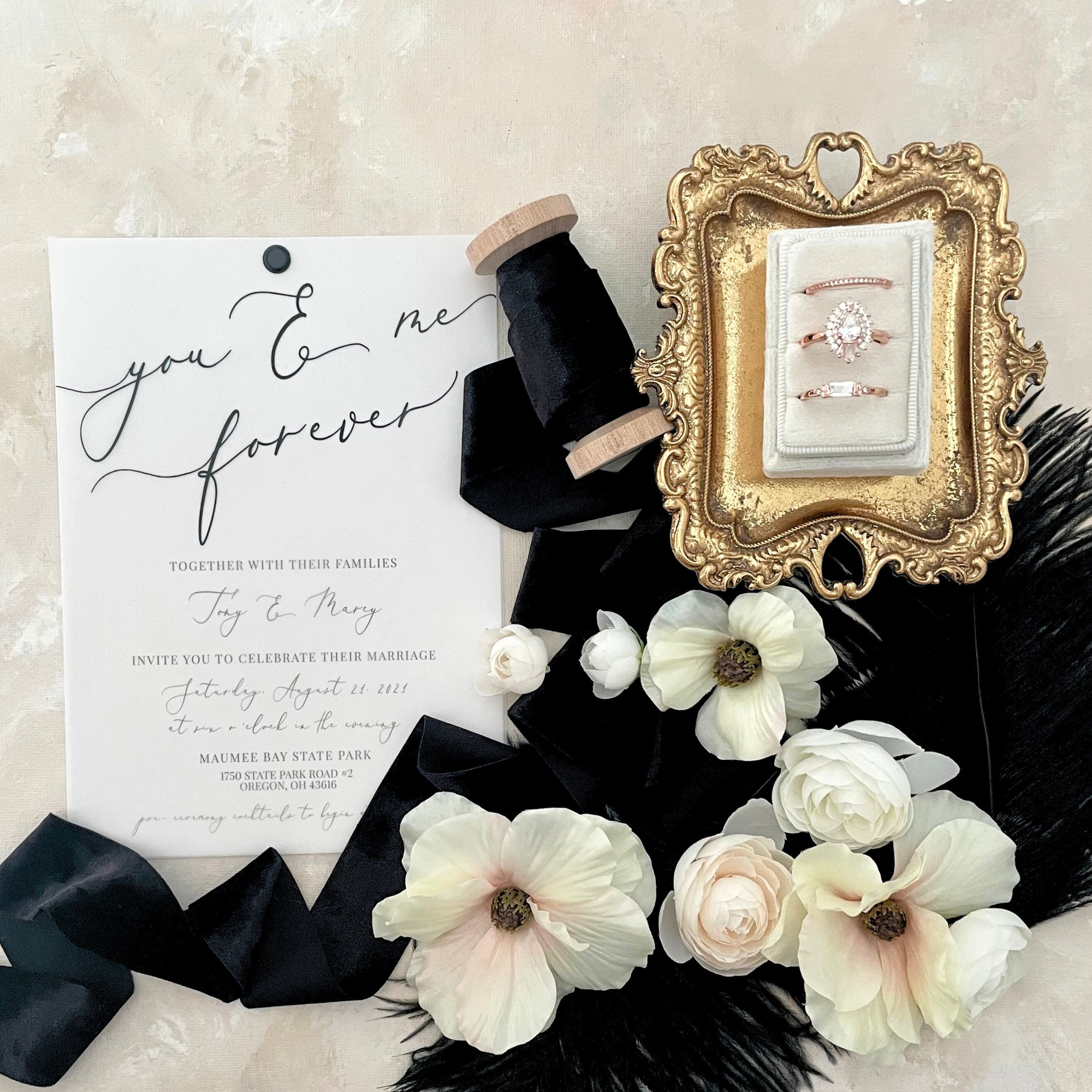 White Wedding invitation with black text.  Black velvet ribbon with mini golden tray with 3 slot white ring box styled with black feathers and white dogwood florals and white roses.