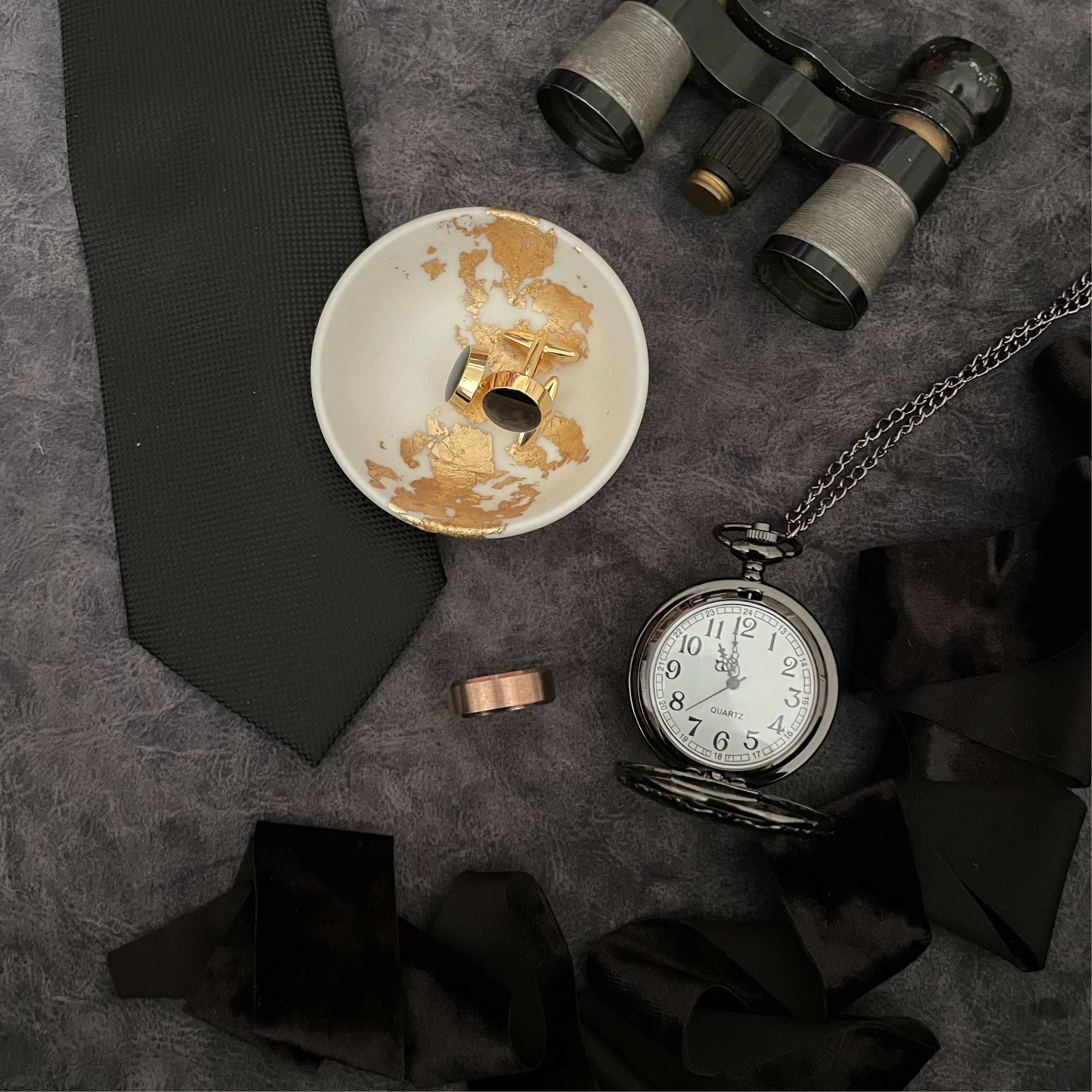 Grooms Flat Lay Detail Styled on dark gray backdrop.  Vintage Silver and Black opera glasses, with black tie, black cuff links & black pocket watch and black velvet ribbon. 