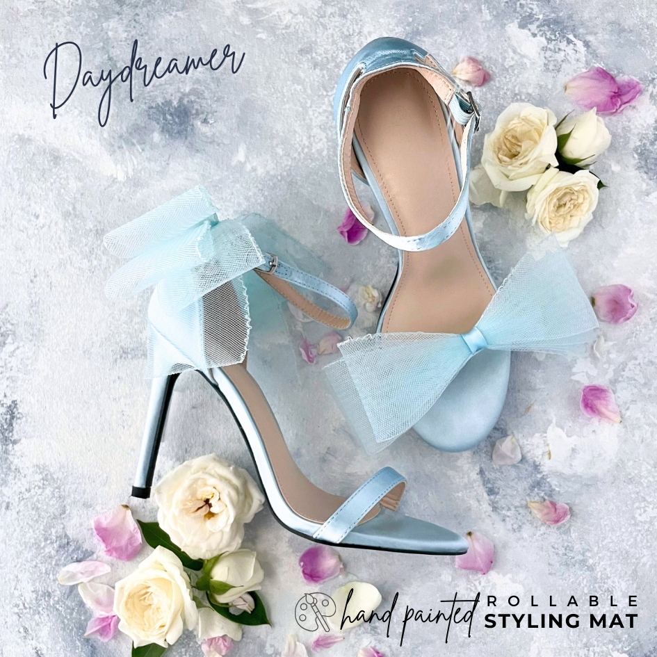 Dusty Blue Bridal Shoe Detail Flat lay Styled on a light blue Hand Painted rollable Flat lay Mat. Shoes are silk light blue with bows, with white roses and pink petals.  Text on image; hand painted rollable Styling mat