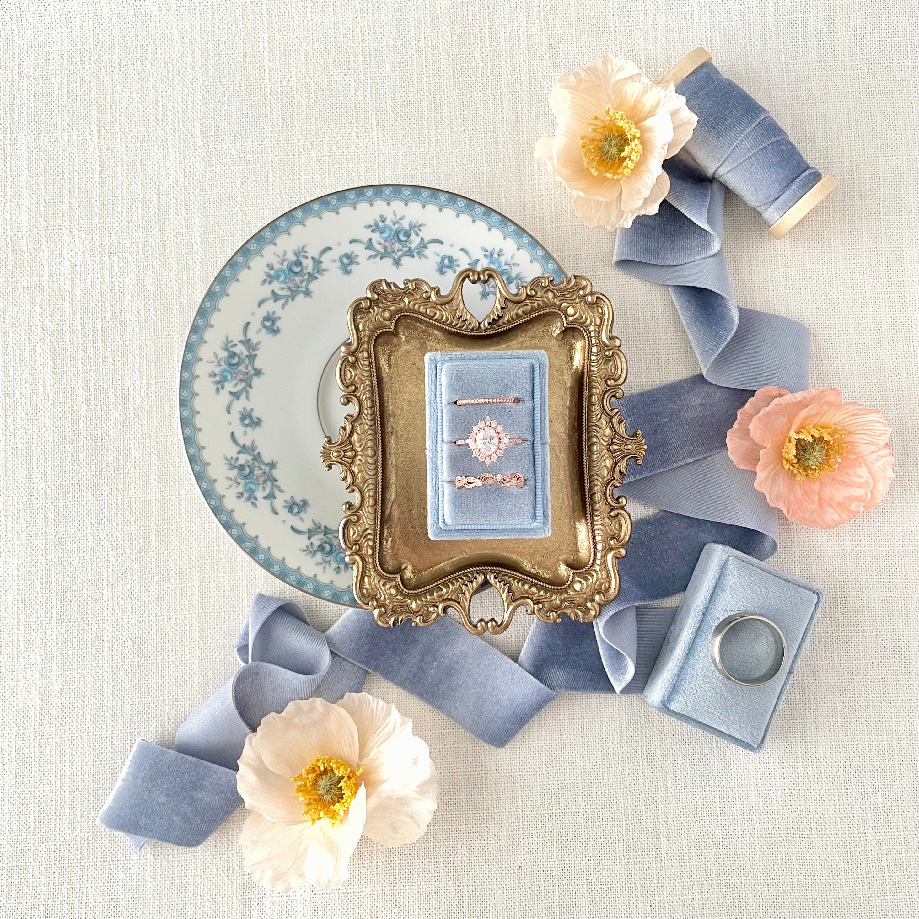 Dusty Blue triple slot ring box on white dish styled with dusty blue ribbon and florals - Wedding Flat lay props from Champagne & GRIT