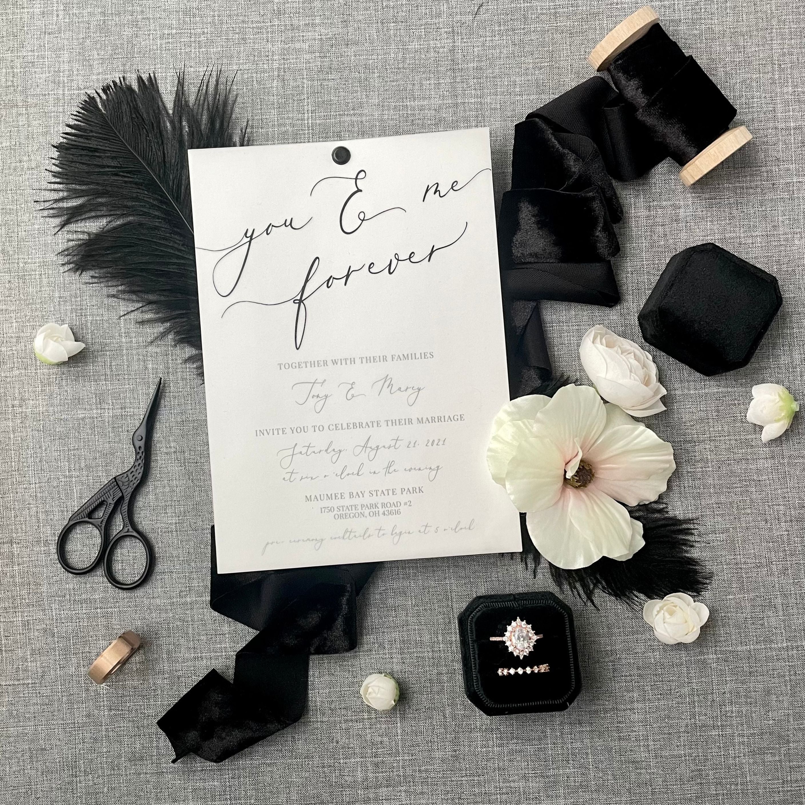 White Invite with Black Scrip styled on a dark gray backdrop with a black ostrich feather, black ring box, black styling scissors and black velvet ribbon.  Wedding Flat lay has white dogwood flowers