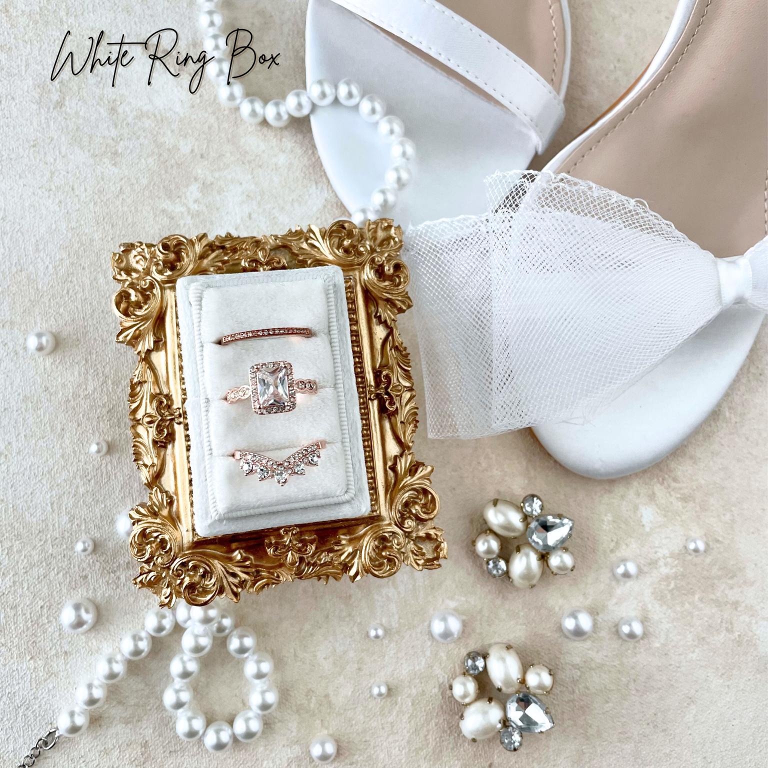 a bridal shoe flat lay detail of a white rectangle ring box, with 3 gold rings.  Styled with a vintage inspired  mini golden frame .  white satin shoes with bows on the strap, a pearl necklace and earrings.  This flat lay is on a tan styling canvas
