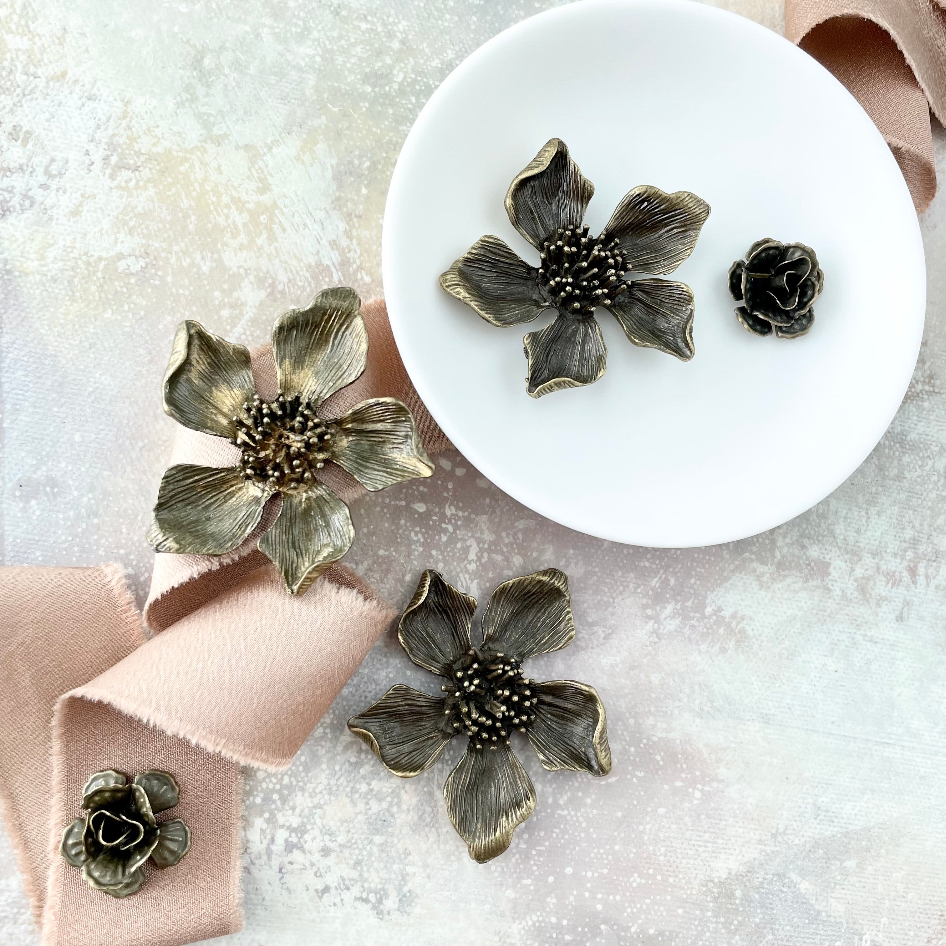 3 large metal styling flowers, two mini bronze styling flowers with a white dish and tan ribbon -  Flat lay props from Champagne & GRIT 
