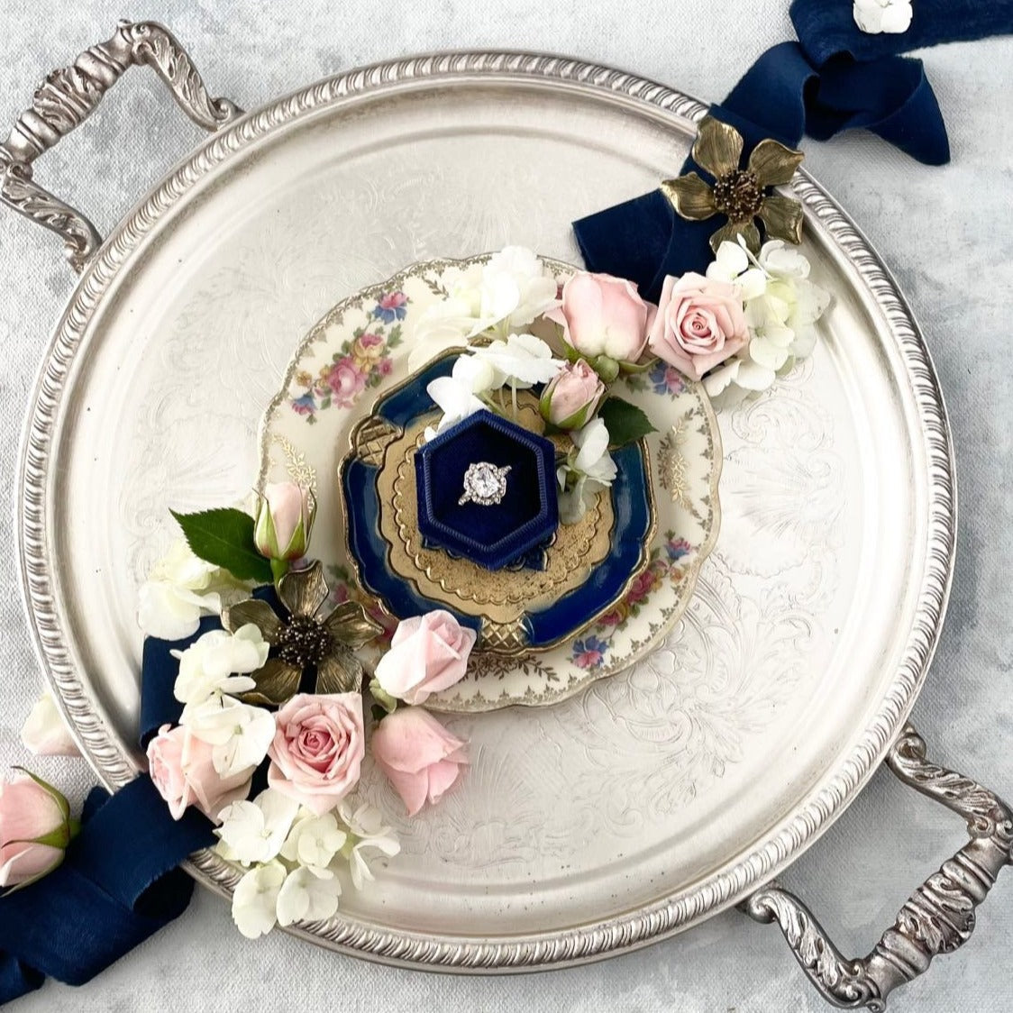 Navy blue ring box on vintage trays styled with navy blue ribbon, white and pink florals and two metal styling flowers  - Flat lay props from Champagne & GRIT