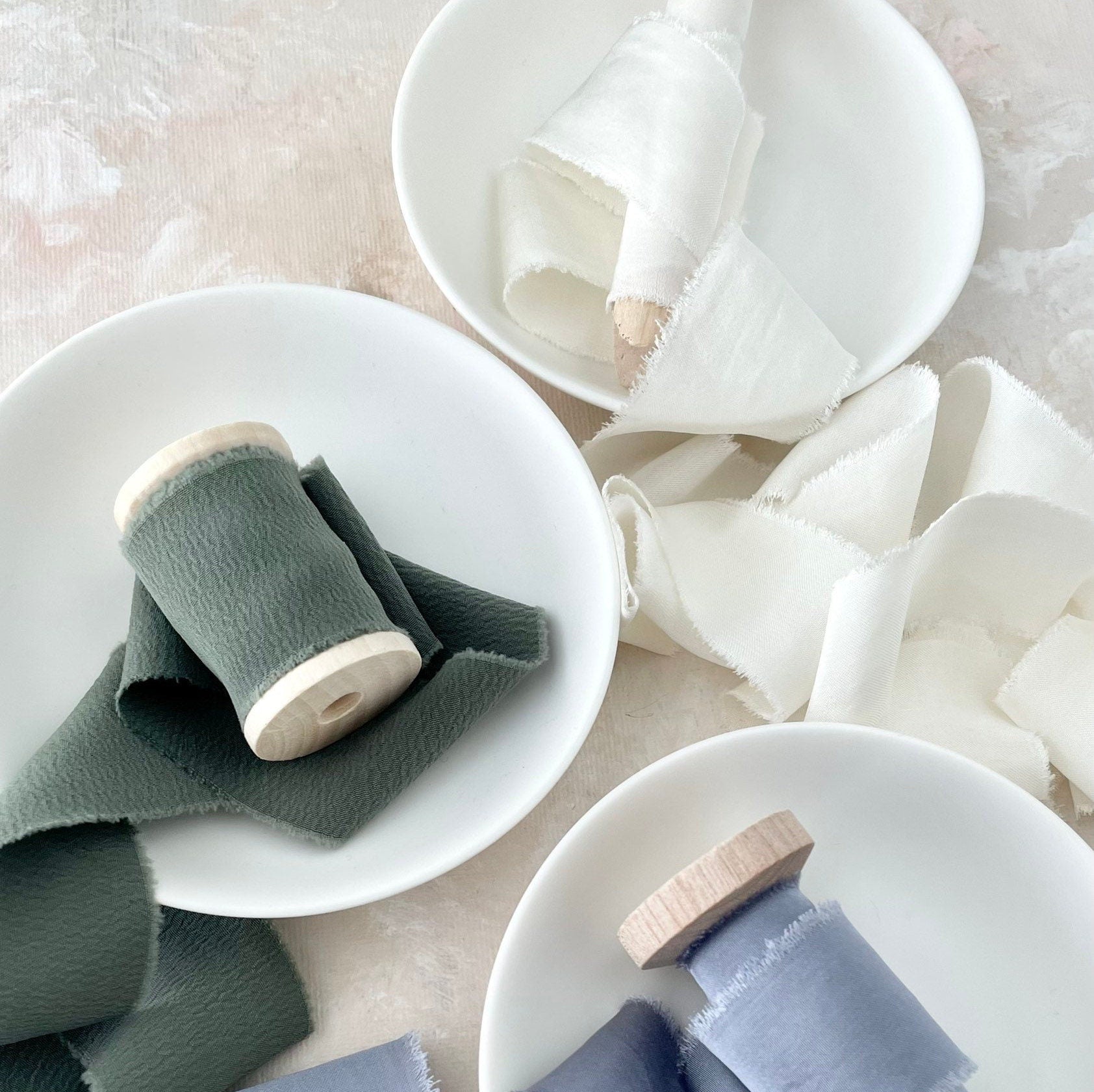 Ivory silk satin ribbon on wood pin styled beside Dusty Blue Ribbon and sage green ribbon in a white tray - flat lay props from Champagne & GRIT