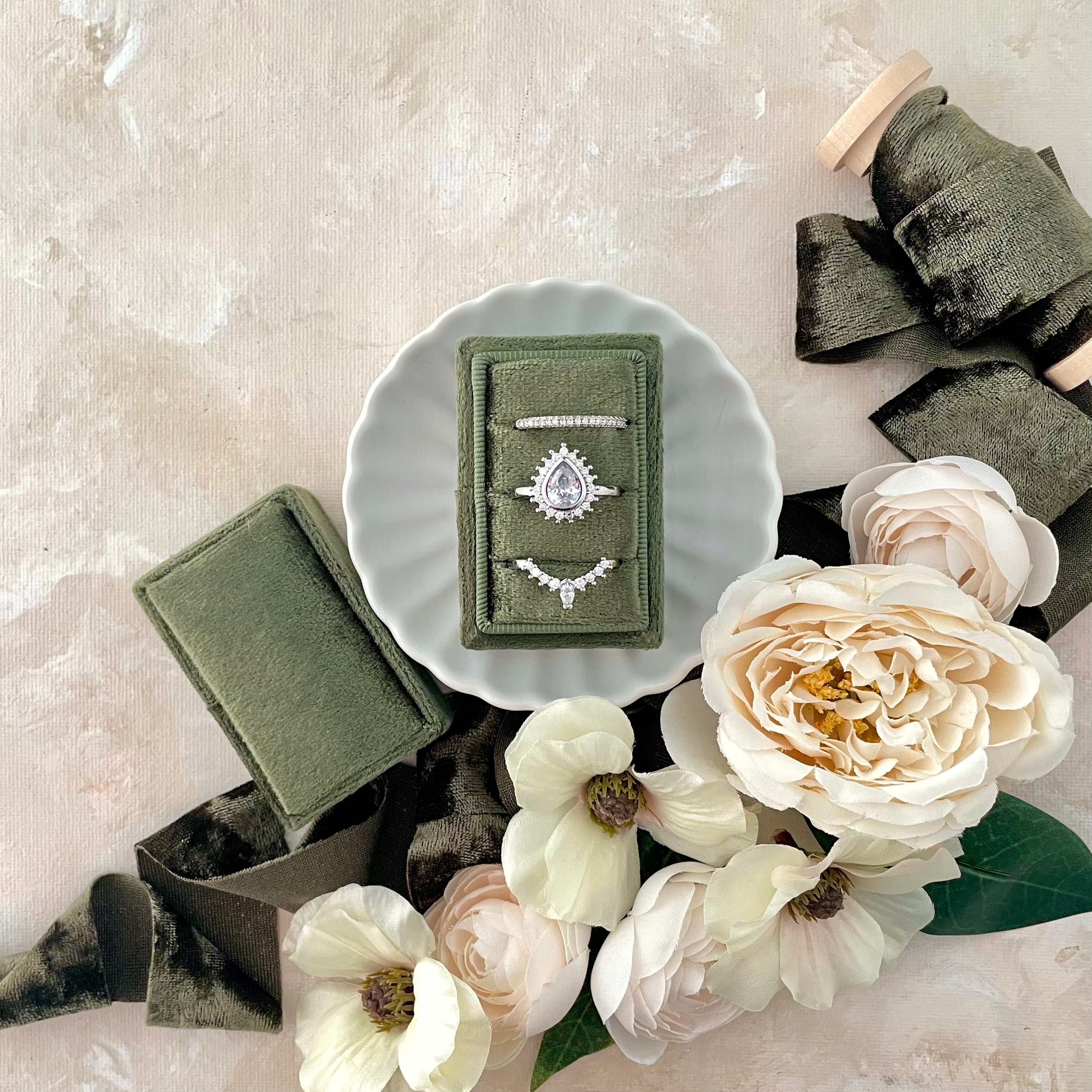 Olive green 3 slot ring box on matte white scalloped dish with olive green ribbon and ivory florals - wedding flat lay props from Champagne & GRIT