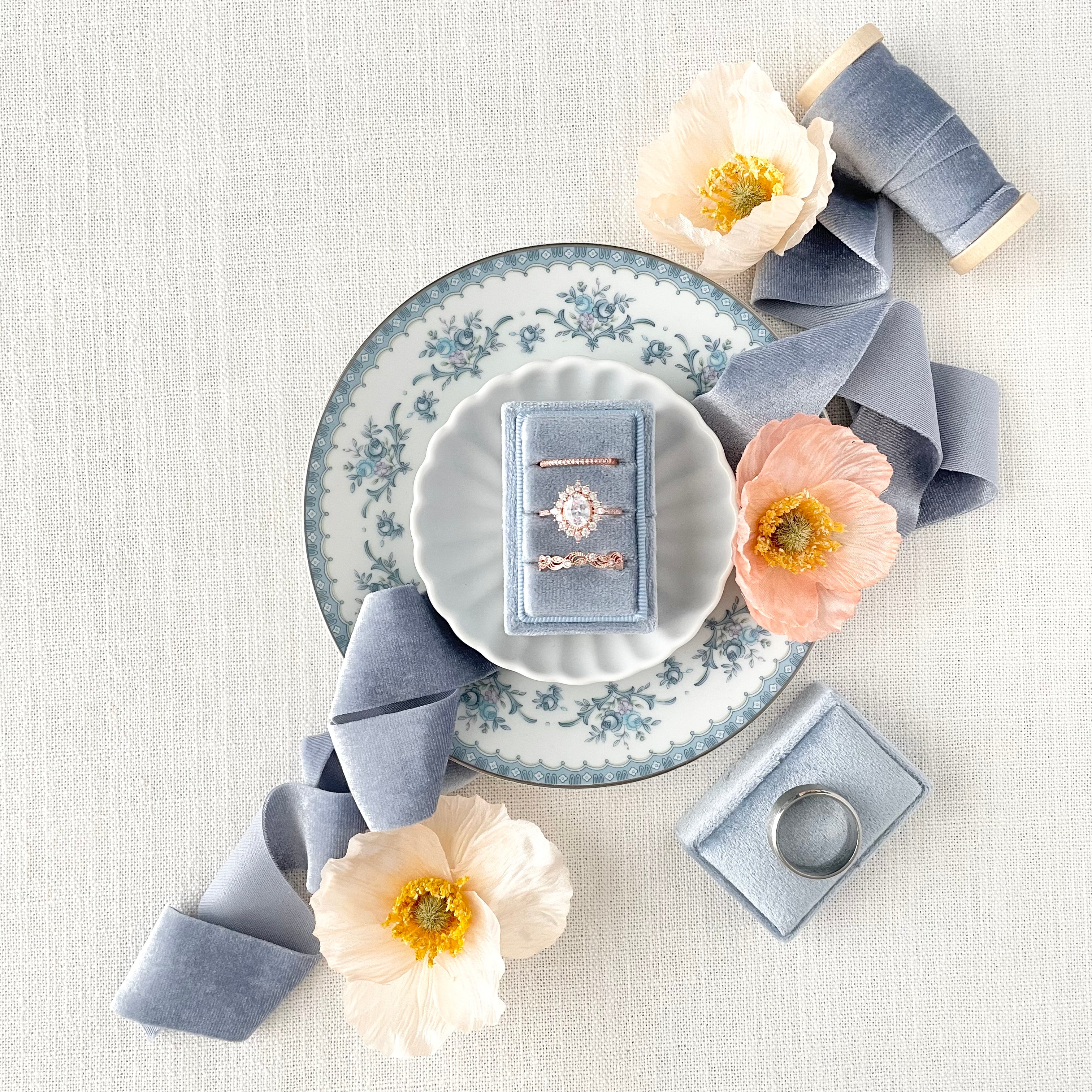 Dusty Blue 3 slot ring box on white dish styled with dusty blue ribbon and florals - Wedding Flat lay props from Champagne & GRIT