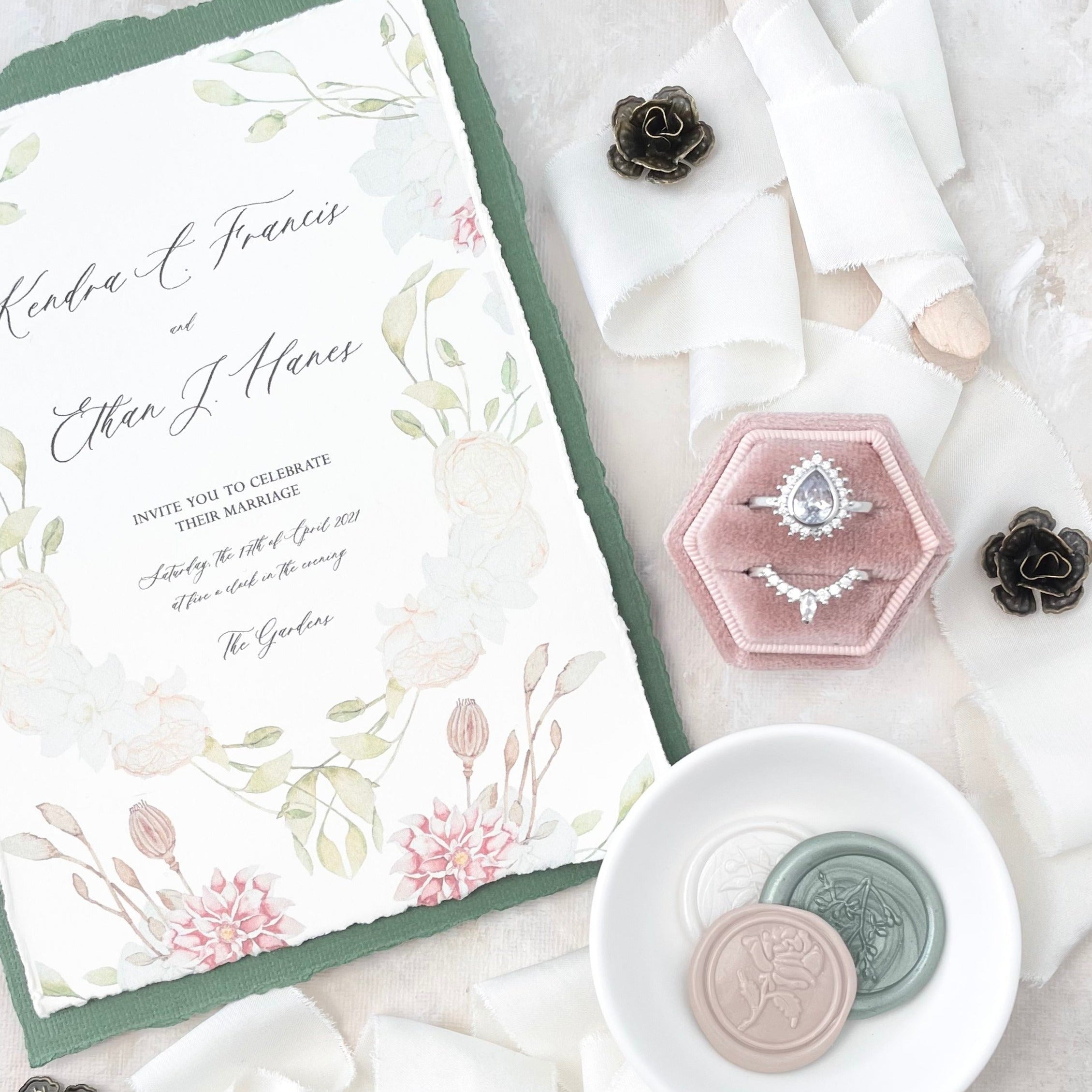 Floral wedding invitation styled with ivory ribbon, 3 wax seals in a white dish, dusty pink velvet ring box, and 2 mini bronze styling flowers -  Flat lay props from Champagne & GRIT 